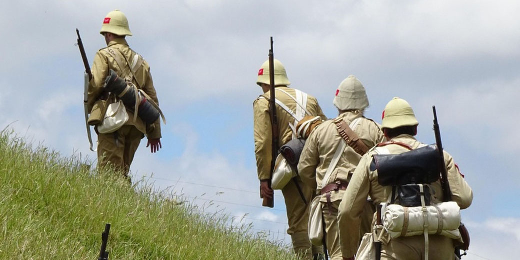Four reenactors in World War I-era Ottoman uniforms march uphill in a line, carrying rifles and field packs under a blue sky.
