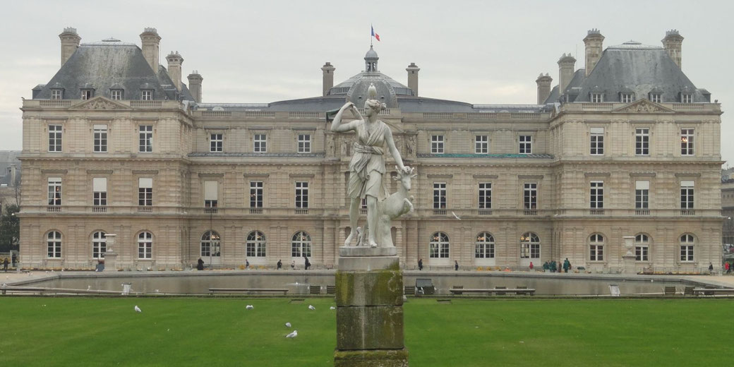A grand historic building with a French flag on top, featuring a central statue of a woman with a deer in a landscaped garden.