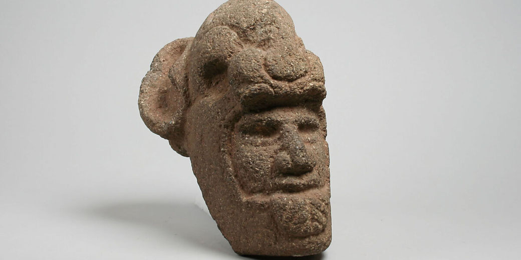 A rough-textured stone sculpture of a human head with a beard, mustache, and an elaborate headdress. The features are stylized and symmetrical, possibly representing an ancient or indigenous figure.
