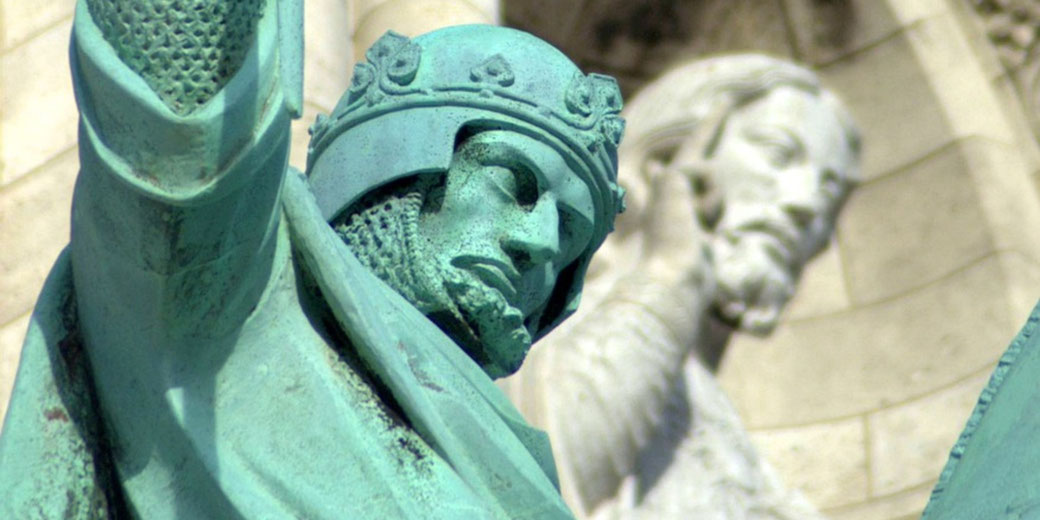 Close-up of a green-patinated statue of a medieval knight in armor, with a stone figure of a bearded man in the background.
