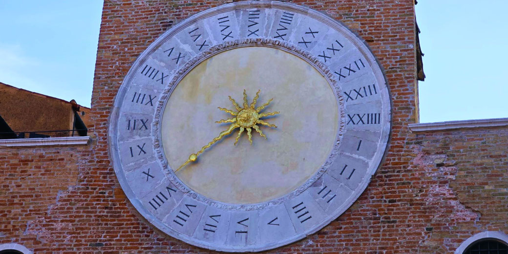 Large stone clock with Roman numerals and a gold sun-shaped hand mounted on a brick tower.