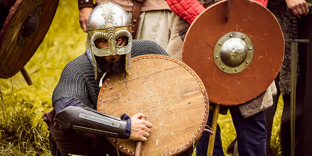A man in Viking armor kneels with a wooden shield and helmet, surrounded by others in medieval gear holding round shields.