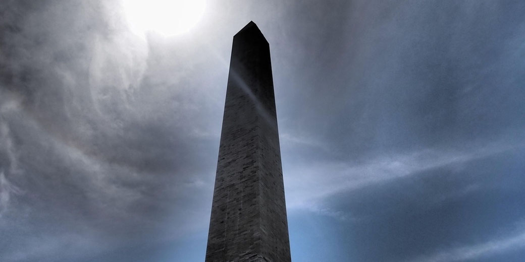 A stone obelisk soars into a cloudy sky, the sun glowing through wisps of cloud above a monumental landmark captured from its base.