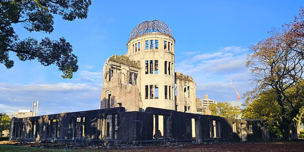 The Atomic Bomb Dome stands in bright daylight, surrounded by autumn trees and framed by a clear blue sky.