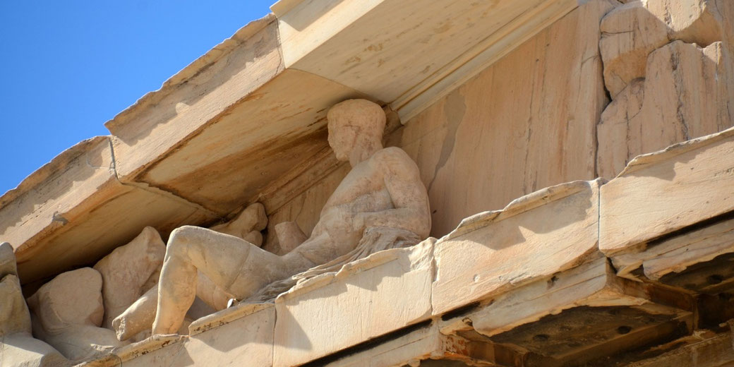 Weathered marble sculptures of reclining male figures on a pediment of an ancient Greek temple, partially damaged, under a bright blue sky.