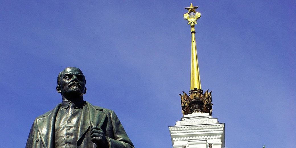 Statue of Lenin stands beside a tall golden Soviet spire topped with a star against a clear blue sky.