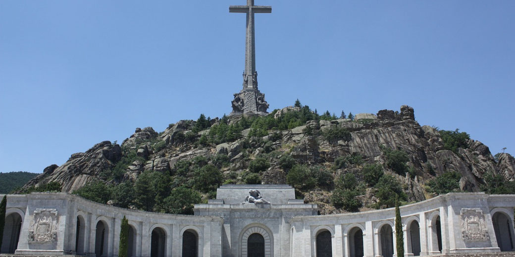 A massive stone cross rises atop a rocky hill above an arched colonnade structure, surrounded by greenery and set against a clear blue sky.