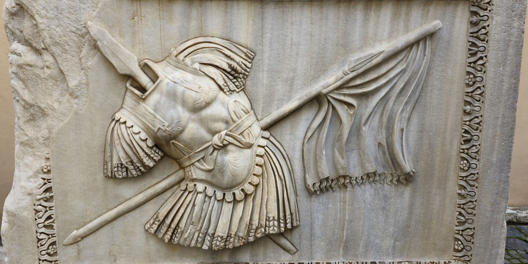 Marble relief depicting Roman armor, a sword, and a flowing flag crossed behind it, all framed by an ornate floral border.