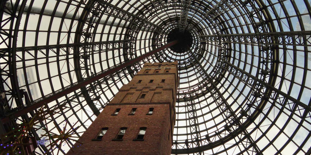 A tall brick tower encased within a large glass and steel cone structure, viewed from below. The geometric dome allows natural light to filter through, creating a striking perspective.