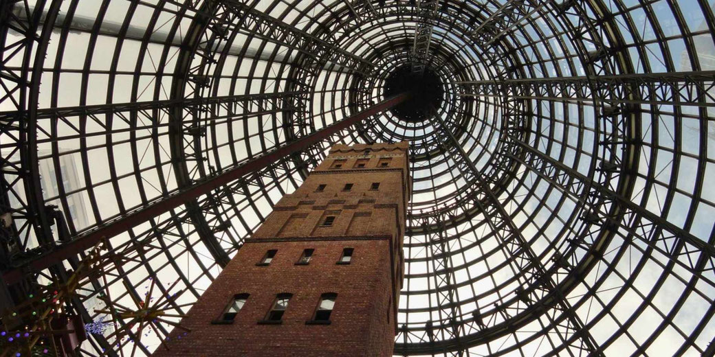A tall brick tower encased within a large glass and steel cone structure, viewed from below. The geometric dome allows natural light to filter through, creating a striking perspective.