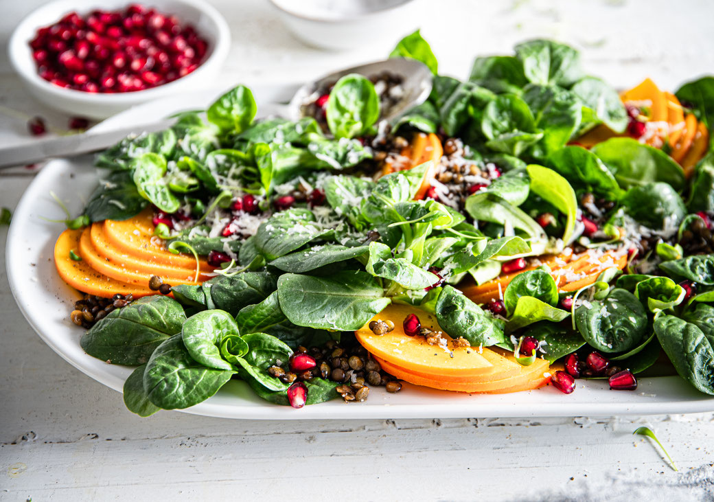 Feldsalat mit Beluga-Linsen, dünn geschnittenen Kaki-Scheiben, Granatapfelkernen und gehobeltem Parmesan auf einer Servierplatte, mit Balsamico-Dressing beträufelt.