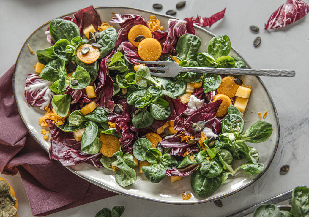 Bunter Herbstsalat mit Kaki, Physalis, Radicchio und Ackersalat, garniert mit gerösteten Kürbiskernen.