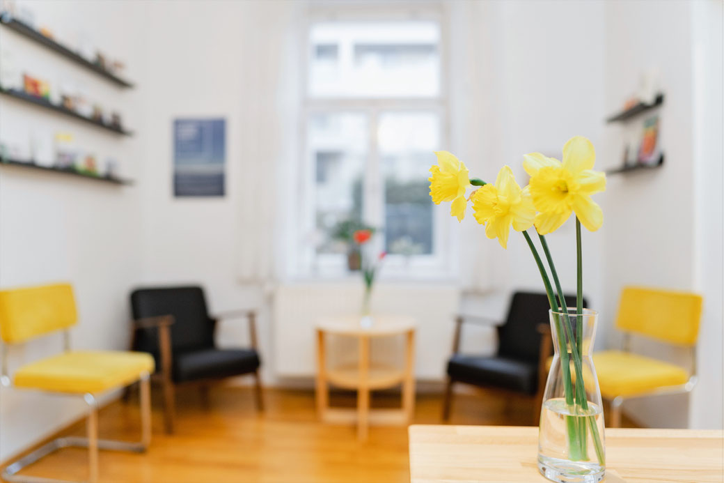 Vor unscharfen schwarzen und gelben Sesseln und einem Fenster in der Mitte kann man scharf 3 gelbe Blumen in einer durchsichtgen Vase sehen.