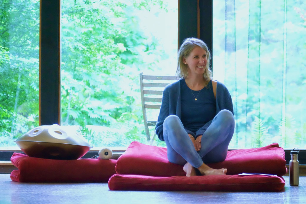 Lächelnde Frau (Maxine Morast) auf Meditationskissen mit goldener Handpan neben ihr im Seminarraum der Grube Louise im Westerwald, dahinter große Fenster mit Ausblick ins Grüne