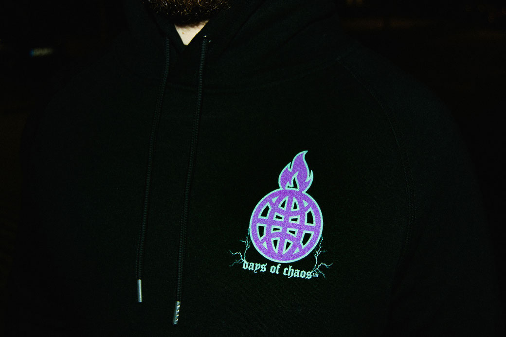 Photo links to the "about" subpage. Shows a close-up of a man at night in a dark environment wearing a black organic metal string hoodie from the underground streetwear brand DAYS OF CHAOS™. The printed logo shows a purple burning globe with lightning.