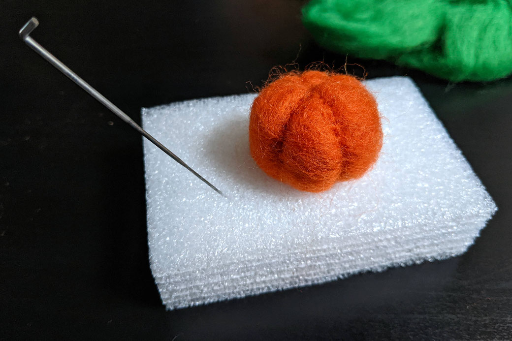 Progress of needle felting a pumpkin shape on foam block, green wool in the background