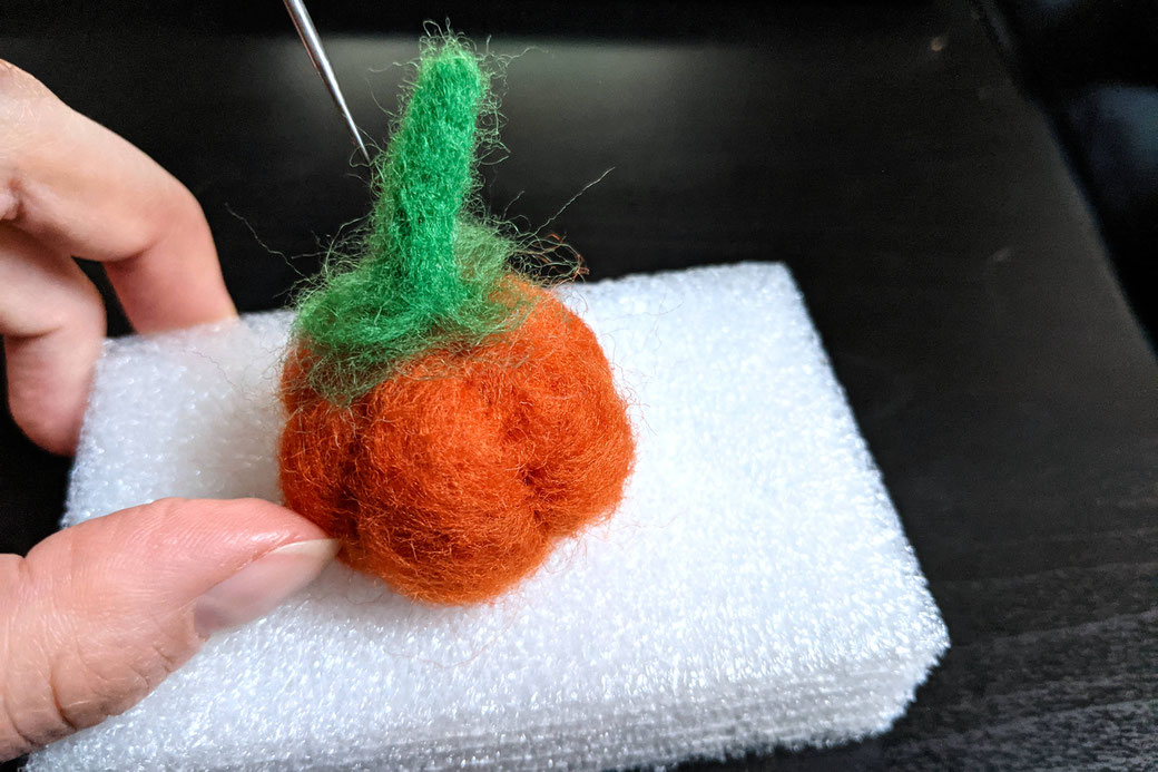 Hand and felting needle attaching green stem to orange pumpkin on foam block
