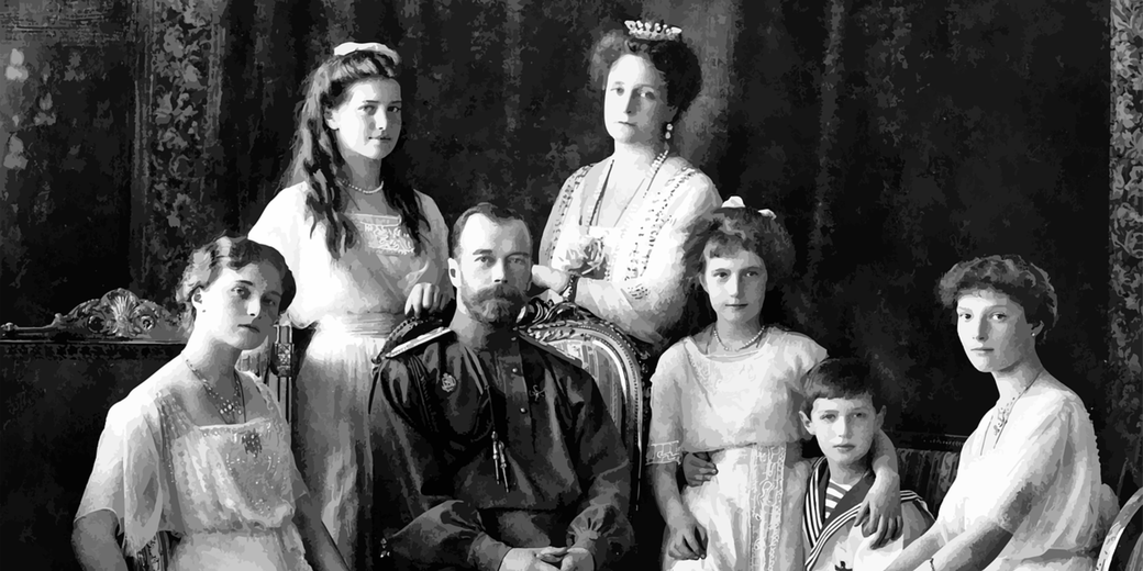 A royal family portrait featuring a man in uniform, a woman in a tiara, and five children dressed formally, all posed indoors.