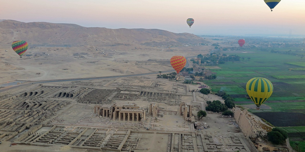 Hot air balloons float over ancient ruins and green fields in Luxor, Egypt, during early morning light with desert hills in the background.
