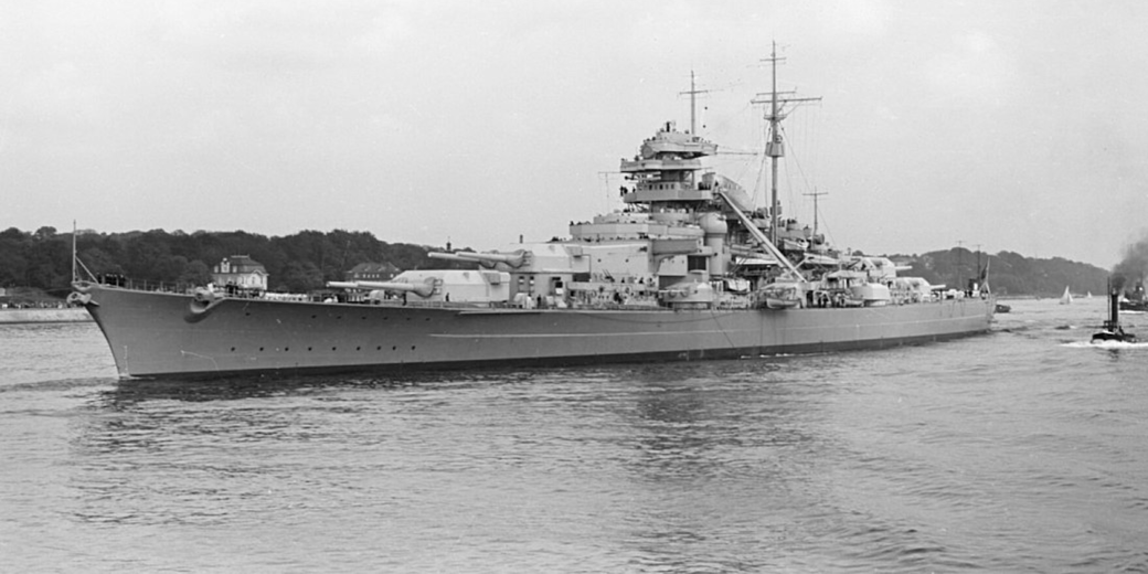 A large battleship with multiple gun turrets moves through a calm harbor near a tree-lined shoreline.