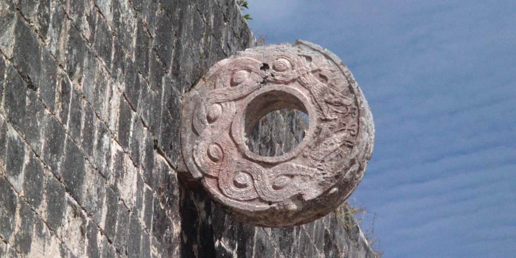 A stone ring with intricate carvings is mounted on a weathered stone wall. It is part of an ancient Mesoamerican ball court, likely from the Mayan civilization, used in ritualistic ball games.