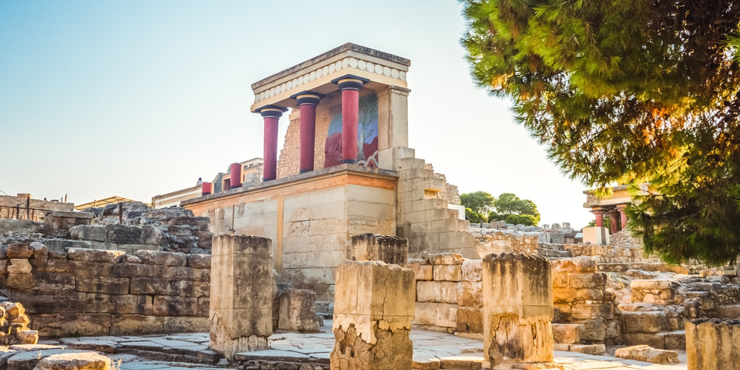 Partially restored ruins of the Palace of Knossos with red columns, stone walls, and surrounding trees in warm sunlight.