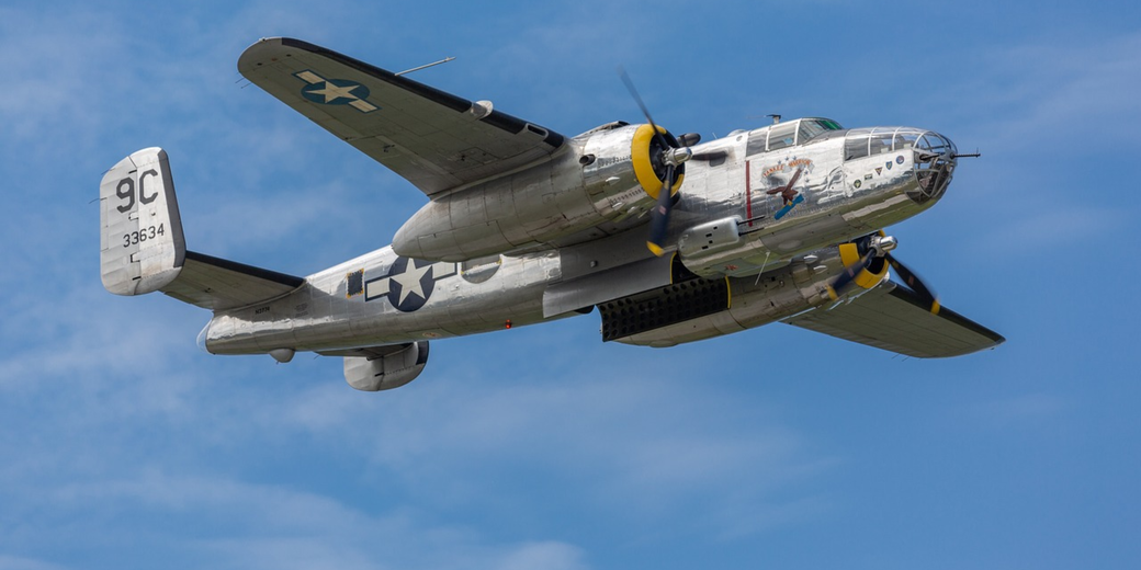 A restored WWII-era B-25 bomber flies midair, displaying U.S. markings and nose art under a clear sky.