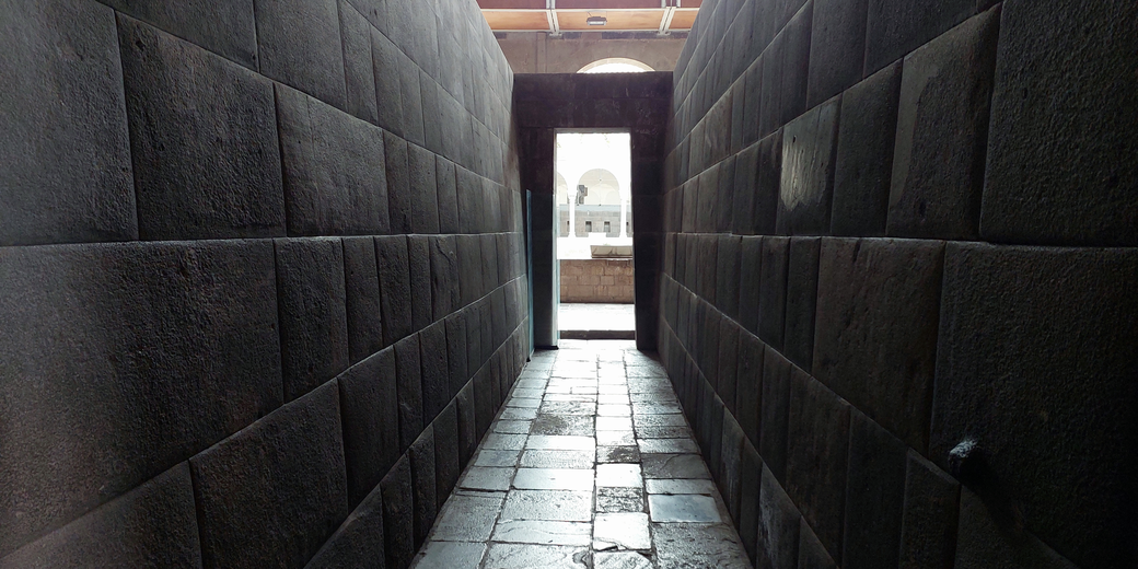 A narrow stone corridor with precisely cut dark stone walls leads to a doorway opening into a brighter courtyard with arched structures in the background. 