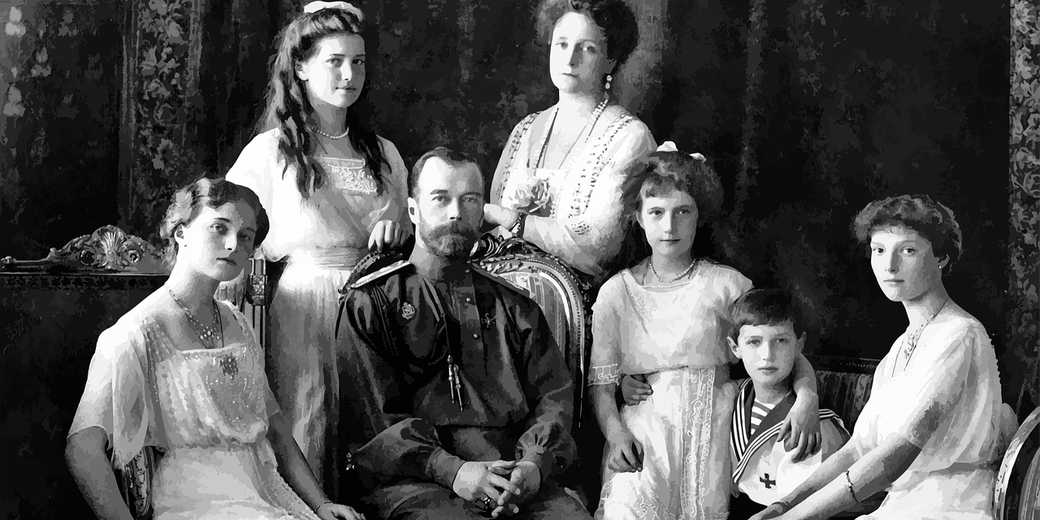 Seven individuals in formal early 20th-century attire pose against a dark backdrop, with the central figure seated and others arranged symmetrically around him.