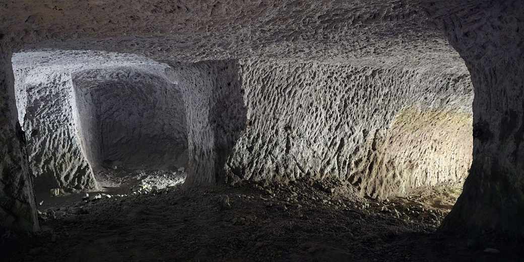 Artificially carved underground tunnel with rough-textured walls and scattered rocks, illuminated by soft directional lighting.