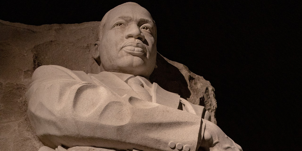 Stone sculpture of a man with arms crossed, gazing forward with a strong expression, carved against a rough rock background at night.