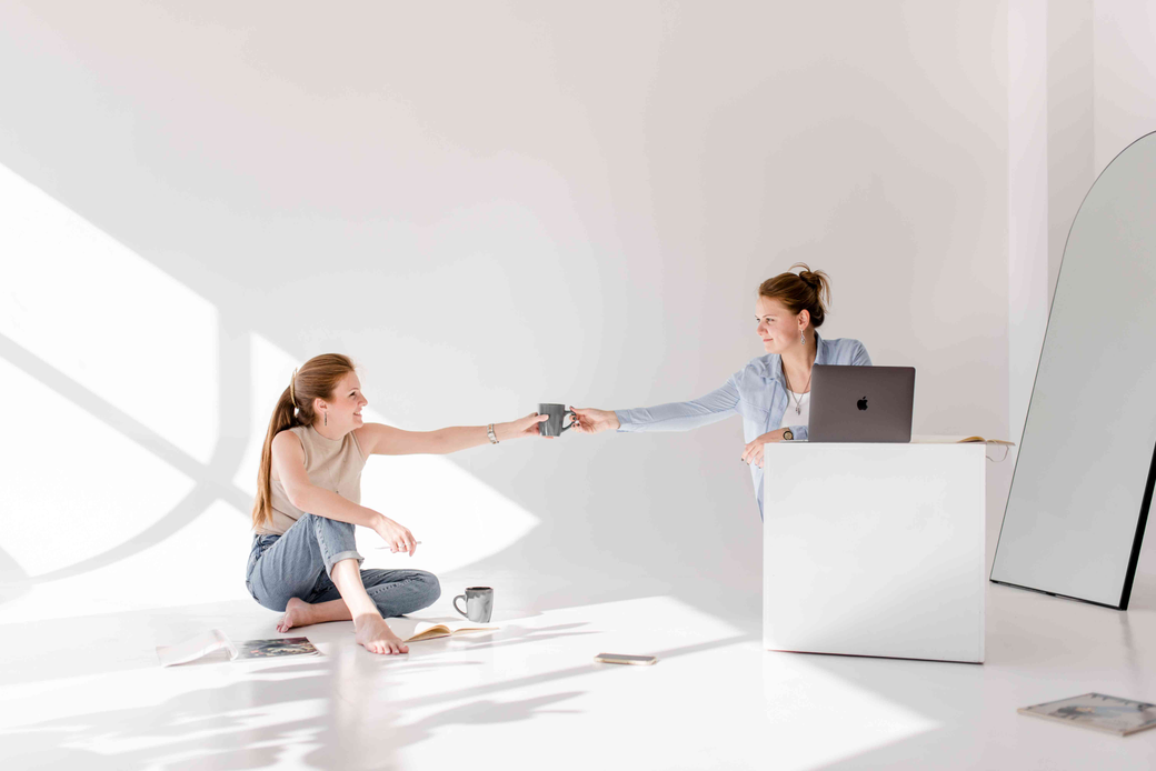 Zwei Frauen die miteinander am Laptop arbeiten, eine reicht der anderen eine Tasse