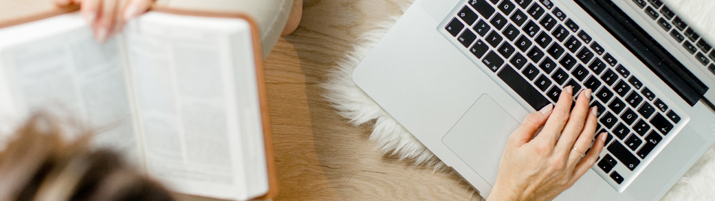 Frauenhände liegen auf der Tastatur des Laptops, die Frau hält ein Buch in der Hand.