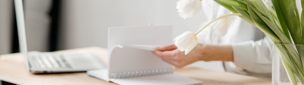 Frau blättert in Notizbuch, Laptop und Blumenvase mit weißen Tulpen stehen neben ihr am Schreibtisch.