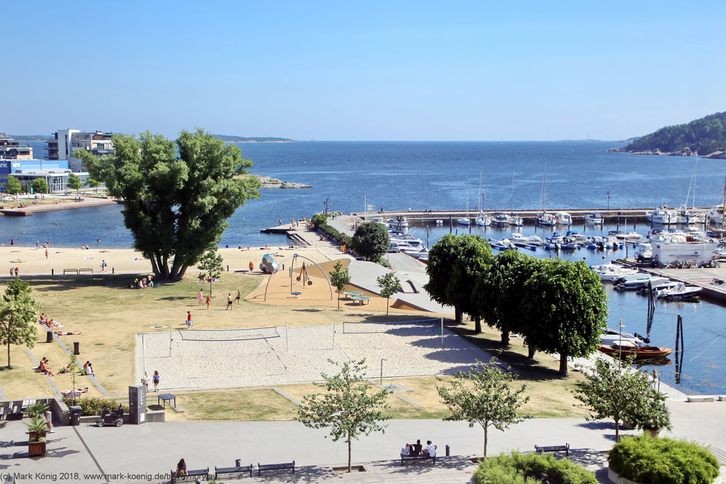 Overview photo of Bystranda Kristiansand showing a beach, voleyball fields, green trees and a yacht harbour in front of the blue North Sea