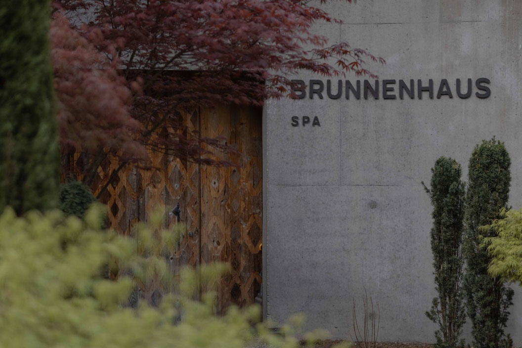 Eingang des „Brunnenhaus Spa“ mit gemusterter Holztür, Sichtbetonfassade und umgebender Gartenlandschaft mit roten und grünen Pflanzen.