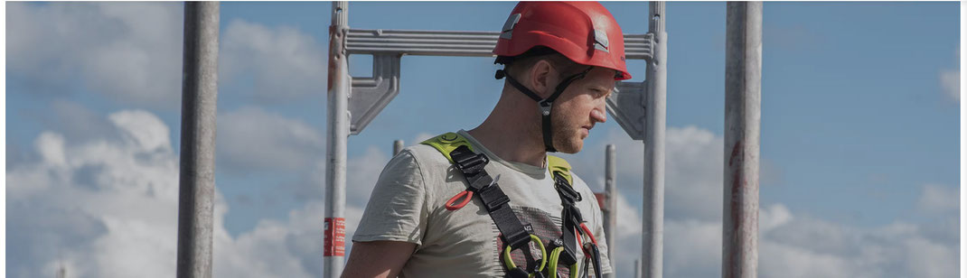 Männliche Person mit einem Arbeitssicherheitsgurt und einem Helm steht auf einem Gerüst und schaut nach unten.