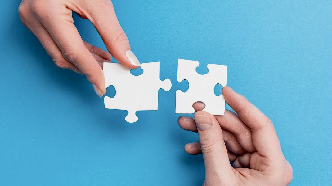 Das Bild zeit die Hände einer Frau und eines Mannes, die beide zueinander passende, weiße Puzzle-Teile in der Hand halten. 