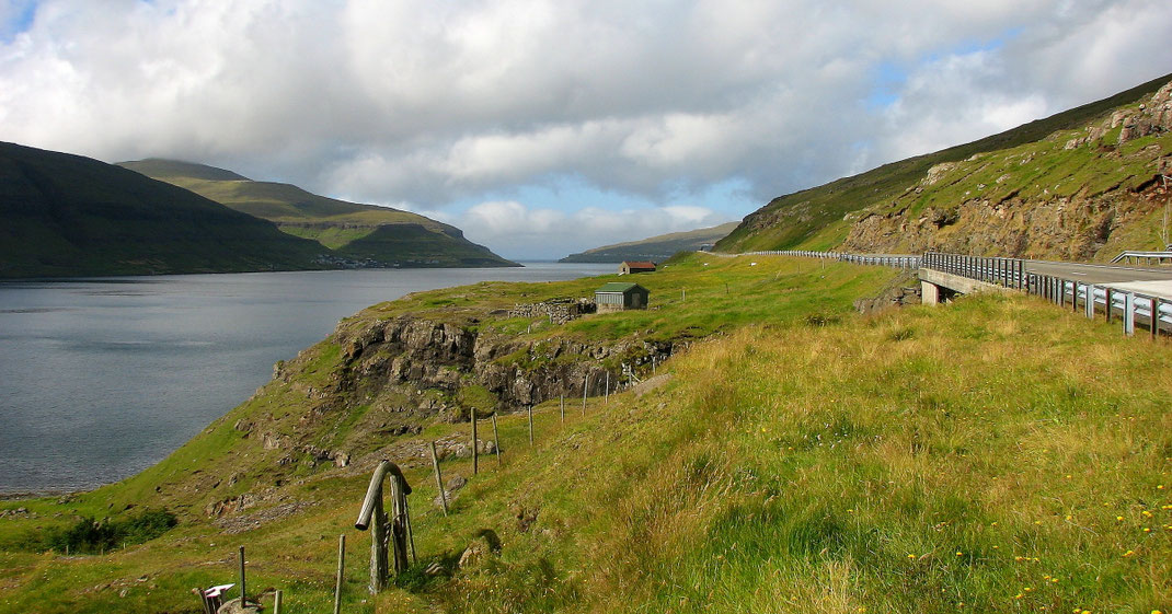 Sehenswürdigkeiten Färöer Inseln: Ein langgezogener Fjord liegt zwischen den bewachsenen Bergen. Eine Landstraße führt entlang des Hangs.