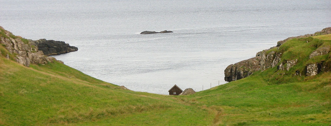 Sehenswürdigkeiten Hoyvík: Eine kleine Fischerhütte steht am Ufer einer Bucht vor Hoyvík. Ein schmaler Fußweg führt durch eine Wiese zur Hütte hin.