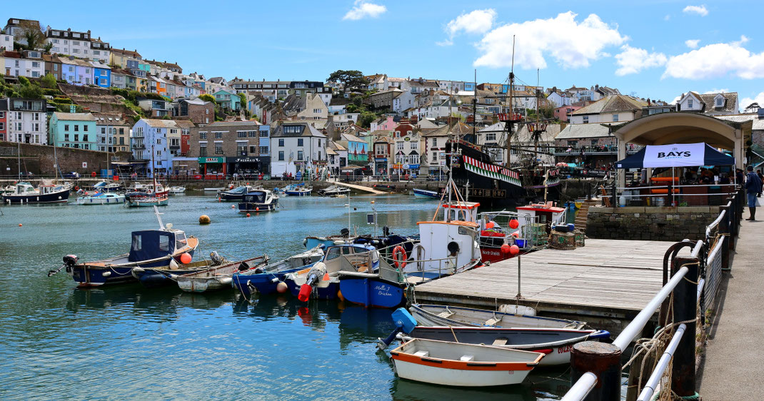 Sehenswürdigkeiten Grafschaft Devon: Bunte Häuser stehen um den alten Fischerhafen von Brixham. Ein alter Dreimaster mit Piratenflagge steht angetäut am Kai.
