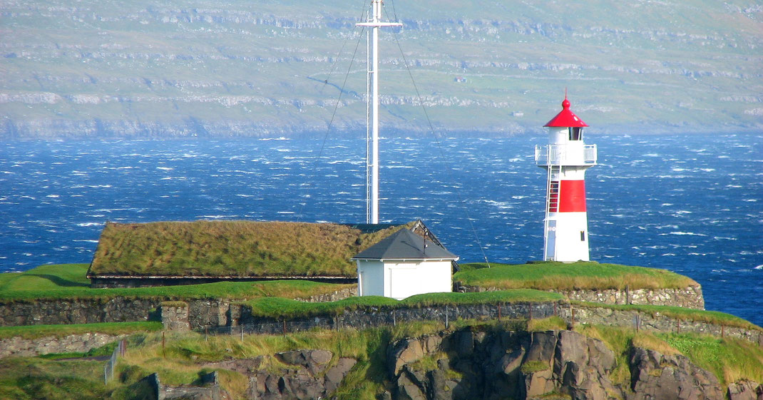 Sehenswürdigkeiten Tórshavn: Der mit roter und weißer Fahne bemalte Leuchtturm steht auf der Festungsmauer der mittelalterlichen Festung Skansin. Ein Steinhaus mit Grasdach steht innerhalb der Mauern.