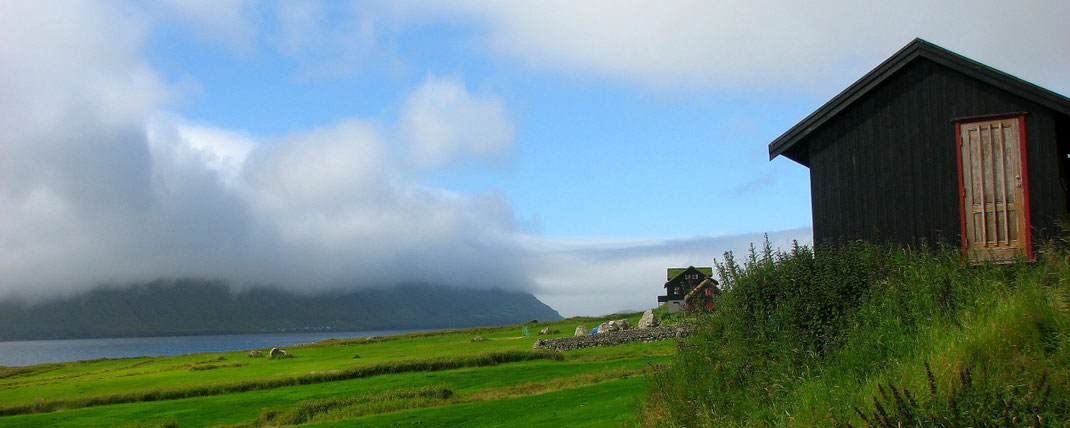 Sehenswürdigkeiten Kirkjubøur: Alte Holzhäuser stehen auf einer sanft zum Meer auslaufenden Wiese. Im Hintergrund ist die mit Wolken verhangene Insel Hestur zu erkennen.
