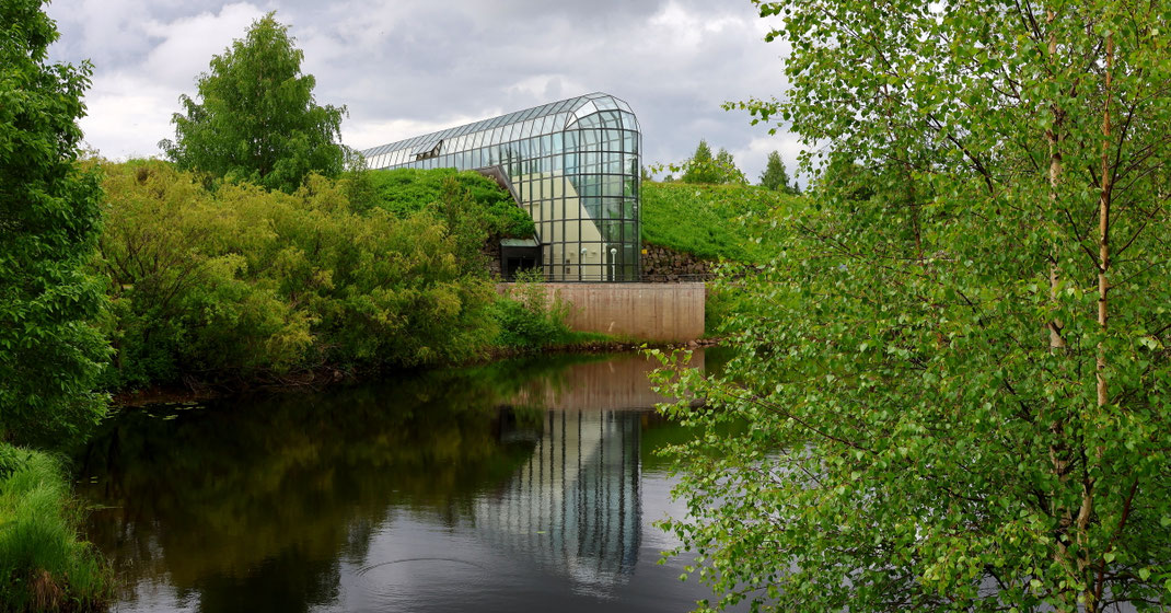 Sehenswürdigkeiten Rovaniemi: Das Glasgewölbe des Arktikum Museums spiegelt sich im kleinen See des Museumsparks. Der See ist von Laub- und Nadelbäumen gesäumt. Ein Fußweg führt entlang des Ufers.