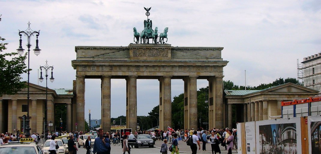 Sehenswürdigkeiten Berlin: Ein emsiges Treiben herrscht um das Brandenburger Tor. Taxis warten mit geöffneten Türen auf Kundschaft, während der Pariser Platz mit Besucher gefühlt ist. Einer der Personen trägt eine britische Fahne, die im Wind flattert.