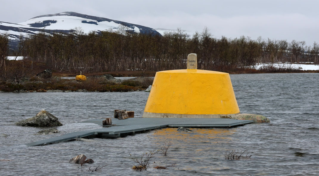 Sehenswürdigkeiten Kilpisjärvi: Der gelbbemalte Grenzstein liegt in einem See. Ein Weg wurde hinüber gebaut, der aufgrund der Schneeschmelze zum Teil unter Wasser steht. Um den See befindet sich ein Birkenwald. Größere Schneefelder liegen am Ufer.