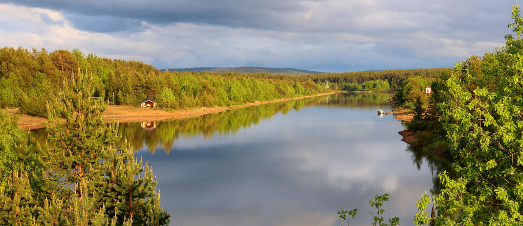 Sehenswürdigkeiten Finnland: Malerisch spiegeln sich die Wolken auf der ruhigen Wasseroberfläche des Vuotso-Kanals. Der Kanal ist gesäumt von Nadel- und Birkenbäumen. Einzelne Hütten stehen am Ufer des Kanals.