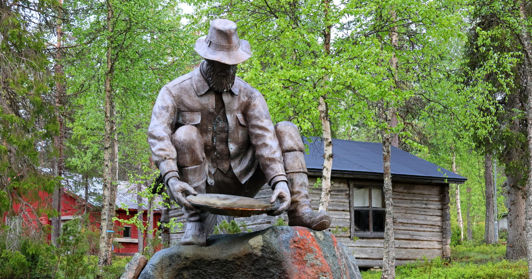 Goldgräberstadt Tankavaara: Bronzestatue eines Goldschürfers. Er trägt einen Hut als Wetterschutz und hohe Stiefel. Er ist in der typischen Hockestellung abgebildet, mit einer Goldwaschpfanne in der Hand, wohl am Ufer eines Baches oder Flusses.