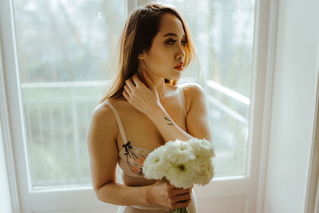 Frau in Dessous mit Blumen in der Hand steht vor einem Fenster