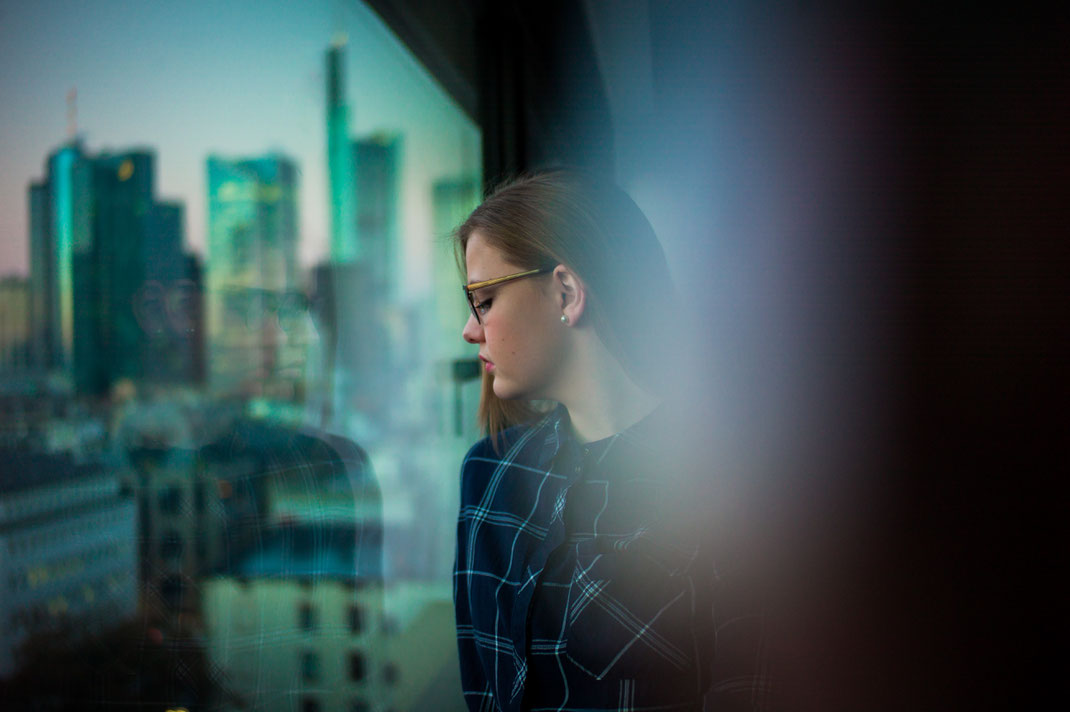 Junge Frau schaut aus Fenster im Hintergrund die Frankfurter Skyline